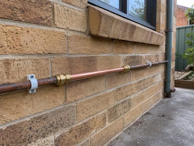 Repaired copper water pipe on a brick exterior wall beside a driveway in Belfield